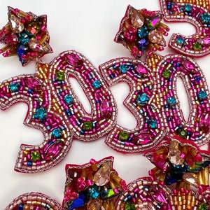 May include: A pair of colorful beaded earrings shaped like the numbers 30. The earrings are made with pink, gold, and blue beads and have a sparkly finish.