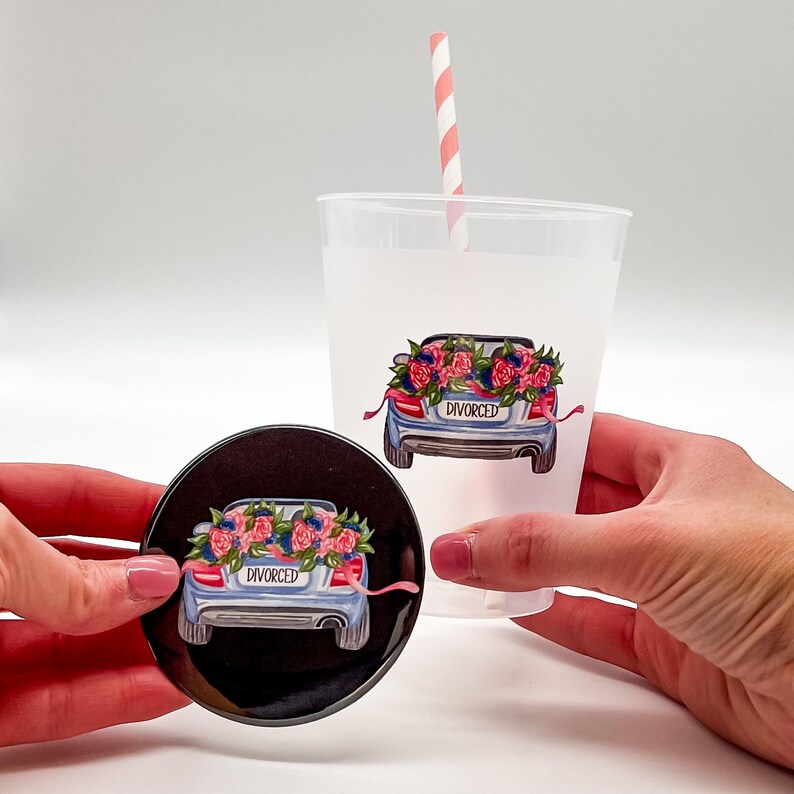 May include: A black round coaster and a clear plastic cup, both featuring a blue car graphic with a floral arrangement and the word "DIVORCED". A pink and white striped straw is in the cup.