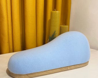 May include: A light blue, padded, curved sewing form with a wooden base. The form is sitting on a white surface in front of a yellow curtain.
