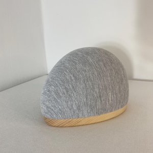 Op de afbeelding: Een grijze stoffen hoed met een afgeronde bovenkant en een licht houten rand. De hoed staat op een lichtgrijze ondergrond. De hoed heeft een eenvoudig, minimalistisch ontwerp.