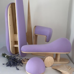 May include: A display of handcrafted wooden items with lavender-coloured upholstery. The collection includes a curved seat, a rounded form, and various wooden components. A bunch of lavender flowers is also visible. The items are arranged on a white surface.
