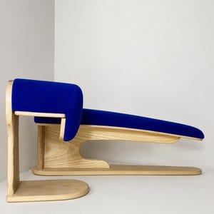Op de afbeelding: Een moderne, sculpturale bank met een blauw gestoffeerde zitting en rugleuning. De bank is gemaakt van lichtgekleurd hout, met een gebogen ontwerp en een laag profiel. De lengte is ongeveer 152 cm.