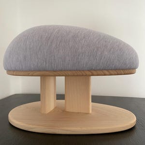 May include: A grey, upholstered hat block with a wooden base. The hat block has a rounded, oval shape and is supported by a light-coloured wooden base with a circular bottom and vertical supports. The hat block is designed for millinery and hat making.