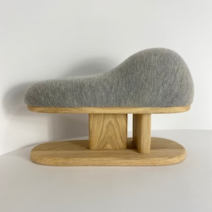 May include: A gray upholstered stool with a unique, wave-like shape. The stool has a wooden base with two rectangular supports.
