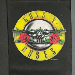 May include: Black fabric patch with a yellow circle containing the words "Guns N' Roses" in a white outline. Inside the circle is a black and white illustration of two crossed pistols with red roses and green leaves. The text "©1988 GUNS N' ROSES" is printed in white at the bottom.