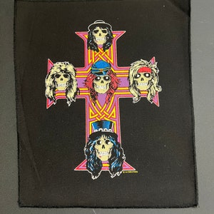 May include: Black fabric patch with a purple and yellow cross and five skull faces of the members of Guns N' Roses. The text "Guns N' Roses" is at the bottom of the patch.