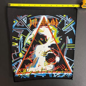Könnte beinhalten: Schwarzer Stoff-Patch mit einem bunten Bild eines schreienden Gesichts in einem Dreieck. Der Text "Def Leppard" und "Hysteria" ist auf dem Bild zu sehen.