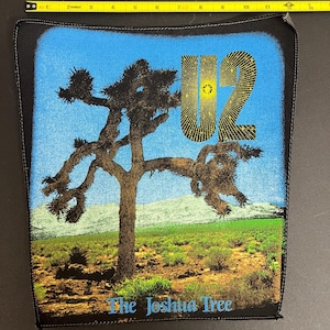 Puede incluir: Un parche de tela negro con un fondo de cielo azul y una silueta de Joshua Tree. El texto "U2" está en amarillo con un efecto de estrella y el texto "The Joshua Tree" está en blanco debajo del árbol.