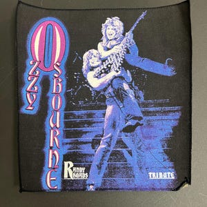 May include: Black and purple patch with a white Ozzy Osbourne logo and a picture of Ozzy Osbourne and Randy Rhoads. The text "Randy Rhoads Tribute" is at the bottom of the patch.