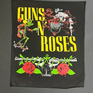 May include: Black patch with the words "Guns N' Roses" in yellow lettering. The patch features a skull with teeth, a skeleton figure, and a gun with roses.
