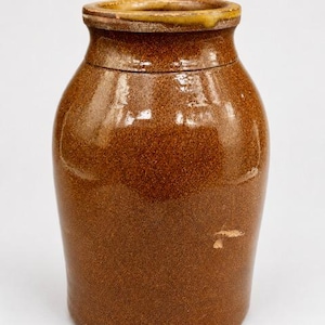 May include: A brown ceramic jar with a wide body and a slightly narrower neck. The jar has a textured surface and a yellow-brown rim. The jar is likely an antique or vintage piece, suitable for kitchen or home decor.