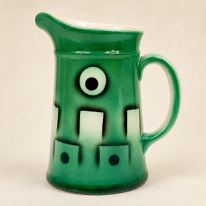 May include: A ceramic pitcher featuring a vibrant green glaze with a white interior. The exterior is decorated with a central black-centered eye motif above a series of white rectangular shapes and small black dots arranged in a repeating pattern.