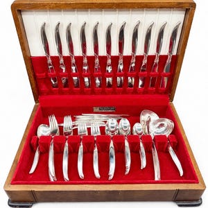 Peut inclure: Un coffret à couverts vintage en bois avec un intérieur en velours rouge. Le coffret contient un ensemble complet d'ustensiles argentés brillants, comprenant des couteaux, des fourchettes, des cuillères et des pièces de service. Les couverts sont soigneusement rangés.