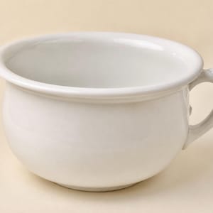May include: A white ceramic chamber pot with a smooth, glossy finish. The vessel features a wide, rounded basin with a thick, rolled rim and a single curved handle attached to the side. The design is simple, functional, and uniform in colour.