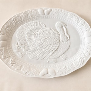 May include: An oval white ceramic serving platter featuring a central embossed turkey figure. The rim is decorated with a raised relief pattern of various fruits, including grapes and pears, set against a smooth, monochromatic white finish.