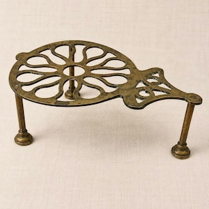 May include: A brass-toned metal trivet featuring a circular top with a cut-out floral petal pattern and an extended handle section. The object is supported by three slender legs with rounded feet, showcasing a weathered, antique-style finish.