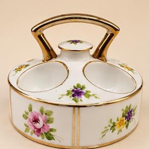 May include: A white porcelain egg holder with two oval indentations and a central handle. The piece features hand-painted floral motifs in pink, purple, and yellow with green leaves. The edges and handle are trimmed with a metallic gold finish.