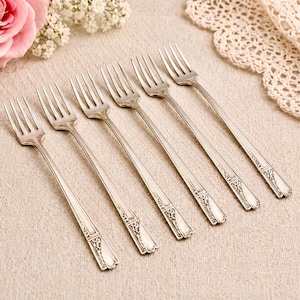 May include: Six silver forks with decorative handles are displayed on a beige linen surface. A pink rose and white flowers are in the upper left, and a lace doily is in the upper right corner.