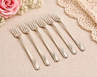 May include: Six silver forks with decorative handles are arranged on a beige linen surface. A pink rose and white flowers are in the upper left corner, and a lace doily is in the upper right corner.