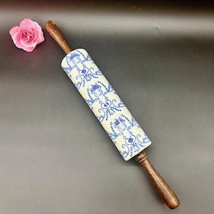 May include: A rolling pin with a white and blue floral pattern. The rolling pin has wooden handles and a ceramic rolling surface. A pink rose is next to the rolling pin.