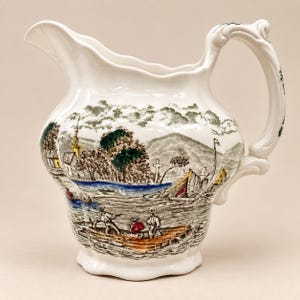 May include: A white ceramic pitcher featuring a detailed transferware scene of a river landscape with figures in a wooden boat, a church, and rolling hills. The design includes accents of green, yellow, blue, and red, with a curved handle.