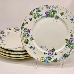May include: A set of four round ceramic plates with scalloped edges and gold-toned rims. The plates feature a white base with a repeating pattern of blue, purple, and yellow flowers with green leaves trailing along the outer border.