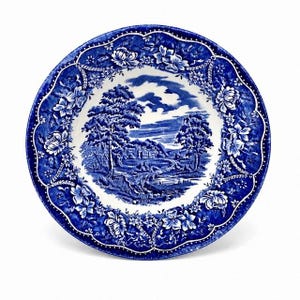 May include: A round, blue and white decorative plate with a landscape scene in the center. The plate features a floral border and a scalloped edge. The landscape scene depicts trees, a building, and a body of water.