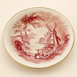May include: A round ceramic bowl with a gold-trimmed rim featuring a detailed red transferware print of a landscape. The scene depicts a windmill, a small house, trees, and figures sitting near a fence, all rendered in a monochromatic red tone.