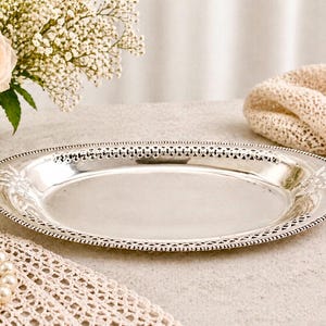May include: A silver oval serving platter with a decorative pierced edge and embossed floral details. The highly reflective platter has a vintage aesthetic. The empty platter is ready for serving. The dimensions are in centimetres.