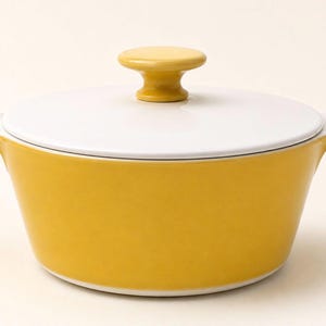 May include: A round ceramic casserole dish with a mustard yellow exterior and a white interior. The dish features two side handles and a matching white lid topped with a mustard yellow knob. The finish is smooth and glossy throughout the piece.