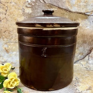 May include: A brown stoneware crock with a lid. The cylindrical container has a dark brown glaze and a lid with a small knob. The number "1" is embossed on the front. The crock is likely used for food storage.