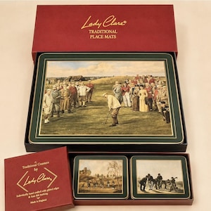 May include: A set of Lady Clare placemats and coasters in burgundy boxes. The placemat features a golf scene with a golfer and spectators. The coasters show outdoor landscape scenes. The text reads Lady Clare Traditional Place Mats.