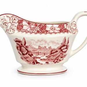 May include: Cream gravy boat with a red floral and landscape design. The rim has a floral pattern, and the body depicts a scenic landscape with a bridge and castle. It has a curved handle and a footed base.