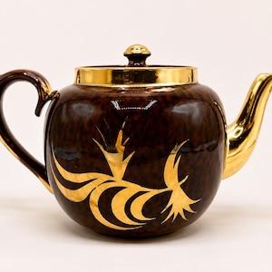 May include: A brown ceramic teapot with gold accents. The teapot features a gold handle, spout, and lid rim. Gold floral designs adorn the body of the teapot, creating a decorative and elegant appearance.