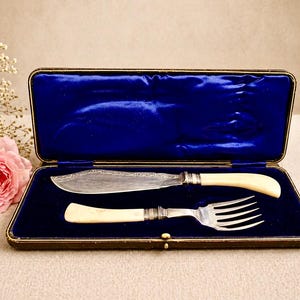 May include: An antique butter knife and fork set in a blue velvet-lined case. The knife and fork have ivory-colored handles and silver-colored metal accents. The case is rectangular with a gold trim.