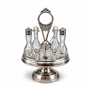 May include: A silver-toned condiment set with six clear glass bottles and jars. The set includes a central jar and five bottles, all held on a tiered, circular stand with a decorative handle. The design is ornate and vintage.