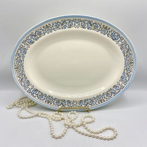May include: A white oval serving platter with a blue and gold floral border. The platter is on a white surface with a string of pearls.