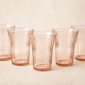 May include: A set of five matching drinking glasses made of translucent pink glass. Each glass features a flared rim and a textured, embossed pattern around the upper exterior, with a smooth base and a starburst design on the bottom.