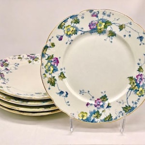 May include: A set of five round ceramic plates with a scalloped edge and a gold-toned rim. The plates feature a white base with a floral pattern consisting of purple and yellow flowers, green leaves, and thin blue stems trailing along the border.