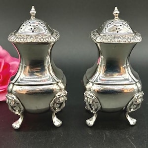 May include: A pair of antique silver salt and pepper shakers. Each shaker has a decorative lid with small holes, a bulbous body, and rests on three small feet. The base of each shaker features a lion's head design.