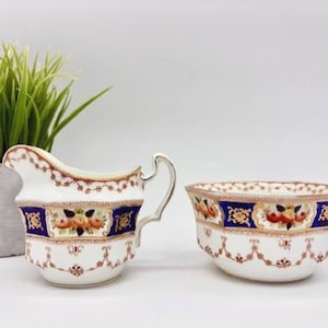 May include: A white porcelain cream jug and sugar bowl set featuring a decorative pattern of dark blue bands, gold trim, and floral motifs in orange and green. The cream jug has a curved handle and a spout, while the sugar bowl is open and round.