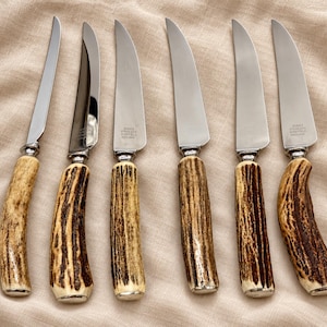May include: A set of six stainless steel knives with textured, brown antler handles. Each blade features a polished silver finish and is stamped with the text JURKS STAINLESS SHEFFIELD ENGLAND. The knives are arranged in a vertical row.