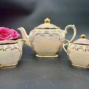 May include: A vintage tea set with a teapot, creamer, and sugar bowl. The set is cream-colored with gold trim and ornate detailing. A pink rose is placed in the creamer. The set is on a dark surface.