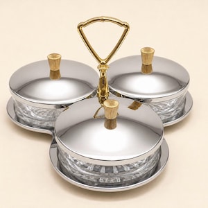 May include: A three-part serving dish set featuring clear glass bowls with textured bases, each topped with a polished silver lid and a small gold-toned knob. A central gold-toned metal handle with a triangular loop connects the three sections.