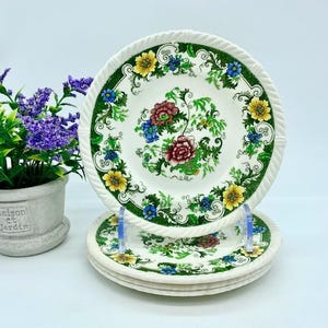 May include: Set of four white ceramic plates with a green, yellow, and blue floral pattern. The plates have a raised edge and a scalloped design.
