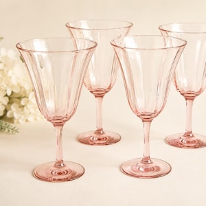 May include: A set of four light pink glass goblets featuring a vertical ribbed pattern on the bowl, a slender stem, and a circular base. The glassware is translucent with a delicate tint, showcasing a consistent design across all four pieces.