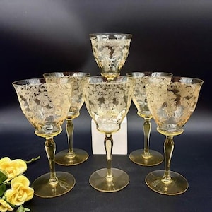 Peut inclure: Ensemble de sept verres à vin ambrés vintage. Chaque verre présente un motif floral gravé sur le bol. Les verres ont une tige fine et un pied rond. Les verres sont disposés sur une surface sombre.