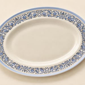 May include: An oval serving platter with a cream-coloured centre and a wide rim featuring a decorative border of blue flowers and intricate dark blue scrolling vines, finished with a solid light blue band along the outer edge of the ceramic dish.