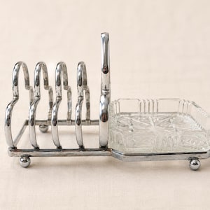 May include: A silver-toned metal toast rack featuring four arched slots for bread slices, attached to a rectangular base with a clear, pressed glass dish with a geometric starburst pattern, all supported by small spherical metal feet.