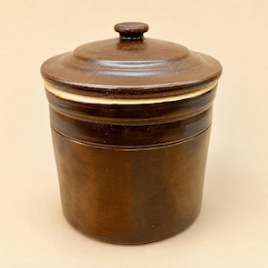 Peut inclure: Un pot en grès cylindrique de couleur marron avec un couvercle assorti. Le récipient présente une finition émaillée marron foncé lisse, un bord légèrement en retrait et un bouton circulaire au centre du couvercle. L'objet est uni.